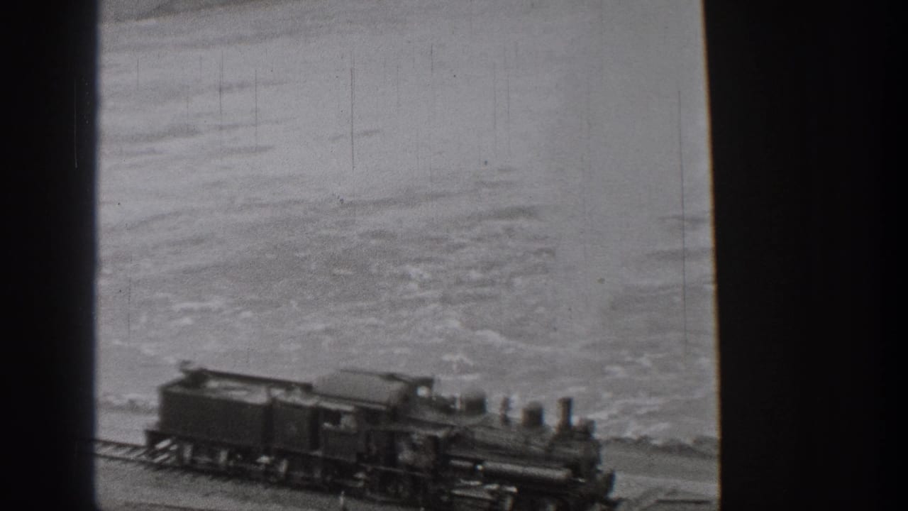 1938 Washington home-movie frame showing a steam train traveling along a coastal track with water and distant hills behind it.