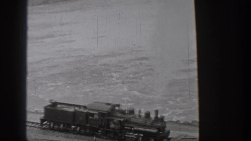 1938 Washington home-movie frame showing a steam train traveling along a coastal track with water and distant hills behind it.