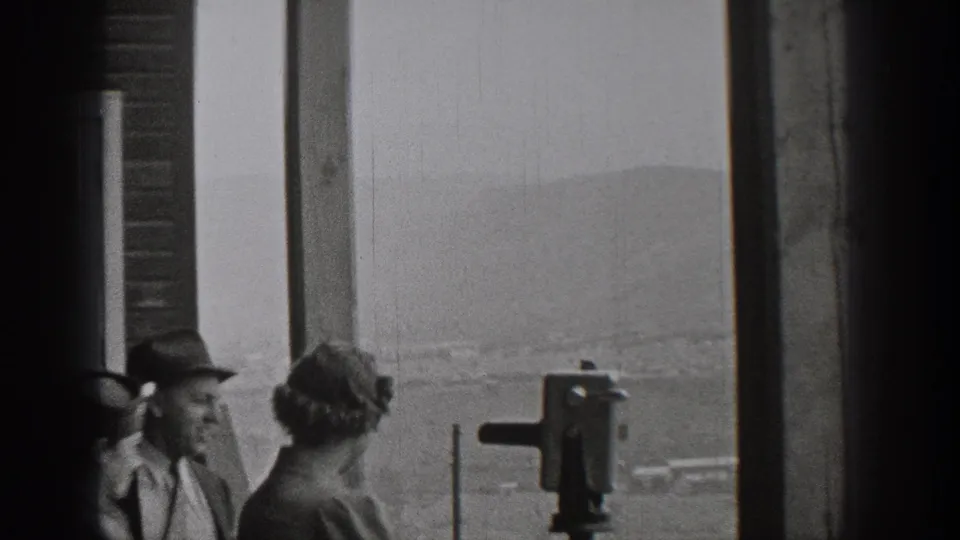 1938 Washington home-movie frame of adults at a scenic overlook, one near a mounted binocular viewer with distant hills behind them.