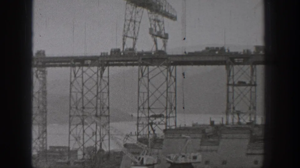 Home-movie view of bridge or waterworks construction in Washington, with cranes set along high steel spans over water.