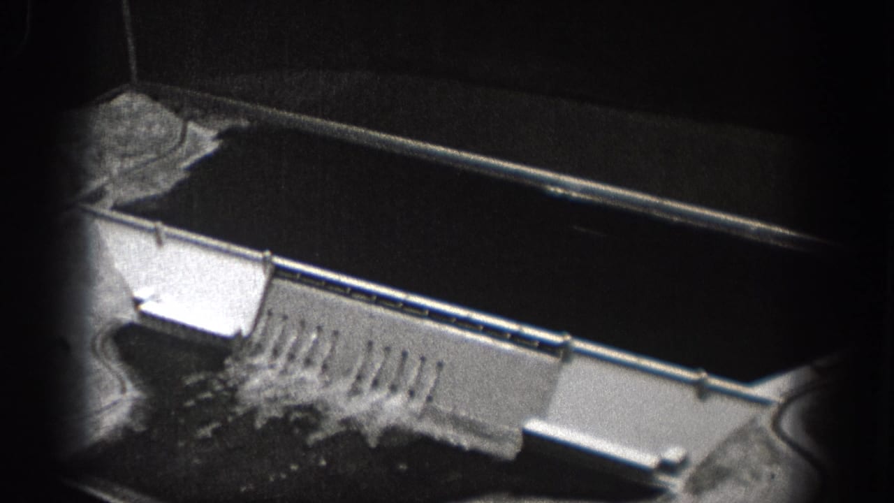1938 Washington 8mm frame showing an aerial view over a dam structure, spillway water, and heavy black film gate edges.