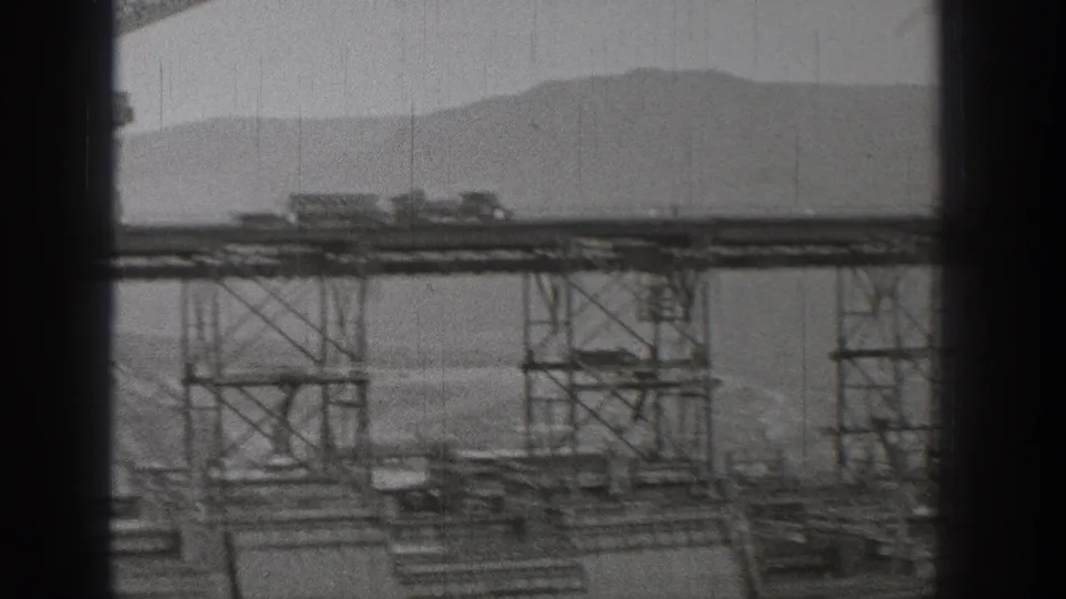 1938 Washington 8mm frame showing distant bridge or dam construction with steel supports, work platforms, mist, and mountain silhouettes.