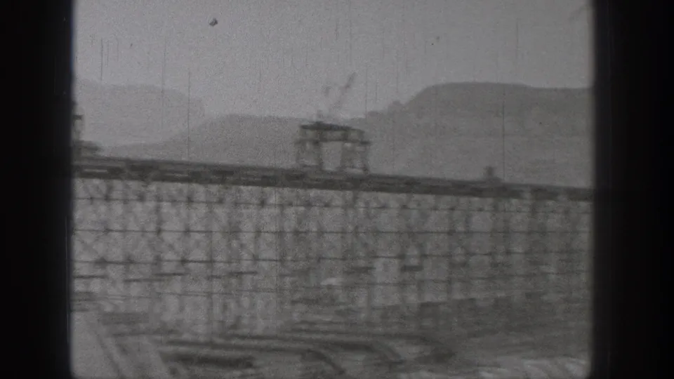 Wide 1938 Washington frame of bridge or dam works, with dense steel trestles, platforms, haze, and a mountain ridge beyond.