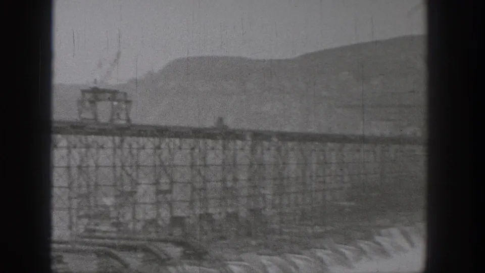 1938 Washington footage appears to show Grand Coulee Dam construction through dark foreground framing, with scaffolding, concrete forms, and hills.