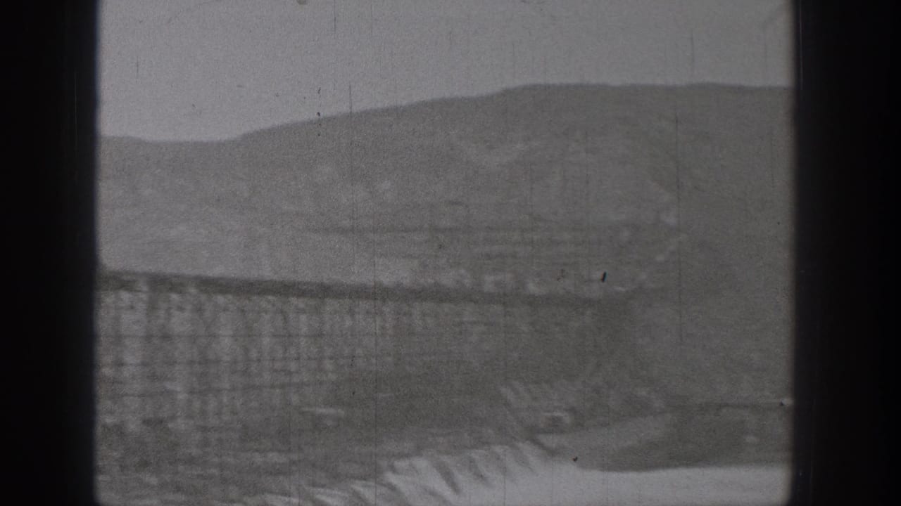 Broad dam structure with scaffolding, spillway-like forms, and hills in the background.