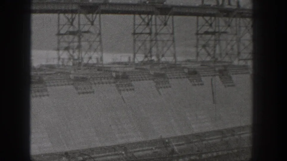 Black-and-white 8mm view of Grand Coulee Dam construction showing lattice supports, concrete surfaces, and a partially obstructed foreground.