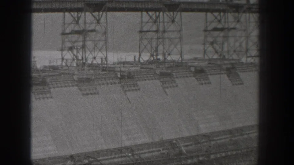 Grand Coulee Dam construction appears in a soft 1938 8mm frame, with scaffold towers, concrete forms, water, and distant hills.