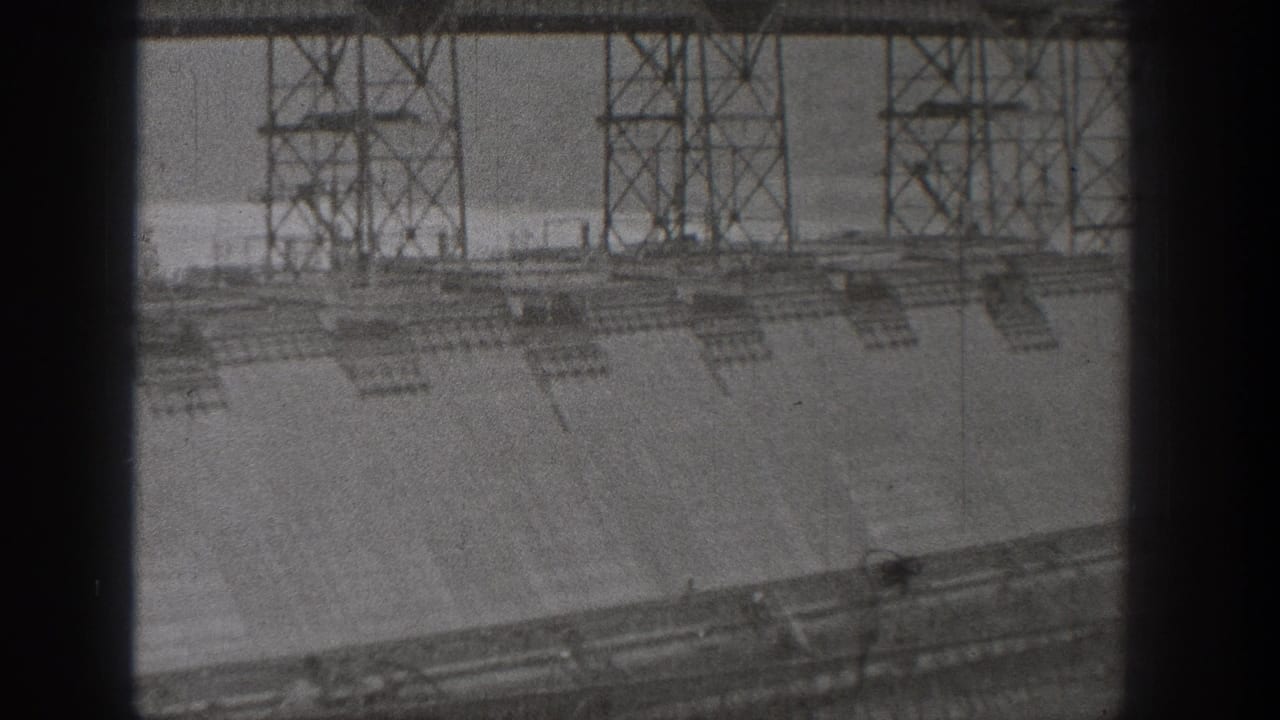 1938 Washington dam construction view with scaffold towers over a concrete wall, seen through a dark 8mm frame edge.