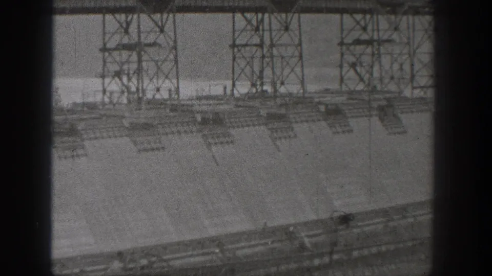 1938 Washington dam construction view with scaffold towers over a concrete wall, seen through a dark 8mm frame edge.