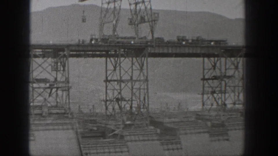 Washington dam construction scene with elevated steel supports, work decking, and hills beyond the site.