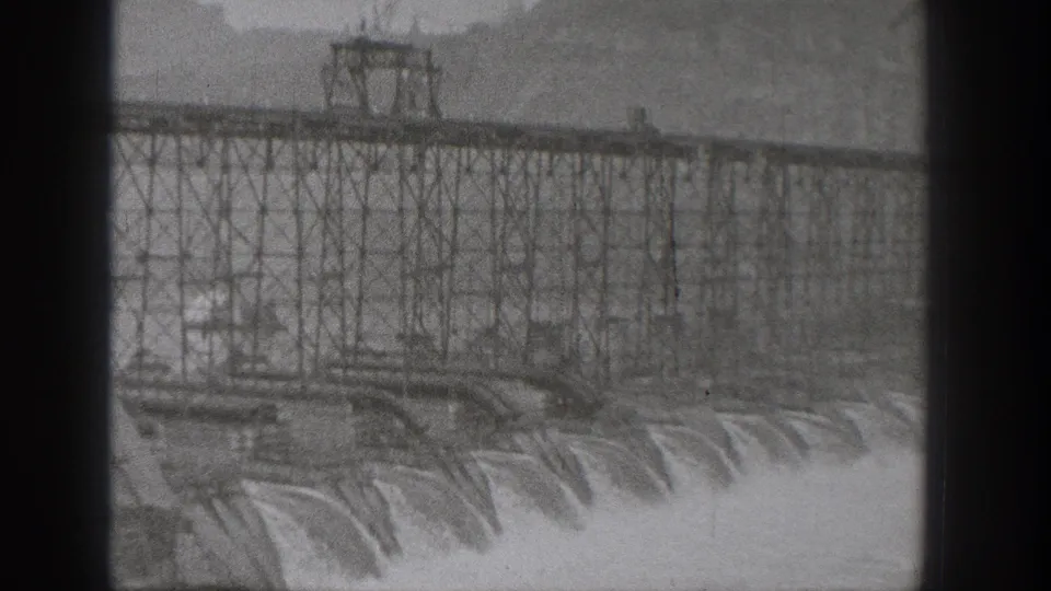 Wide 8mm view of a Washington dam under construction, with dense scaffolding and water rushing below.