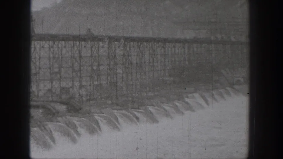 Distant 1938 Washington dam construction view showing steel framing above a line of rushing spillway water.