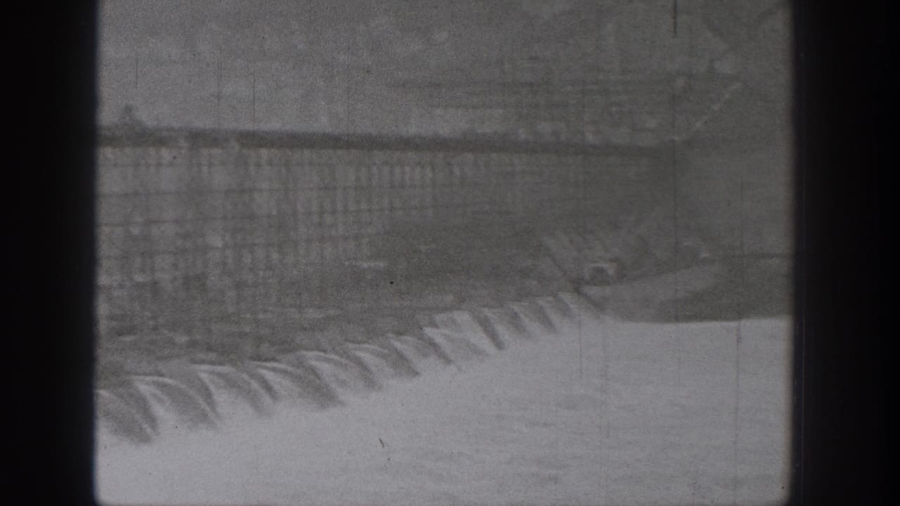 1938 Washington dam footage showing spillway water over concrete, with distant hillside and dark vertical obstruction in the foreground.