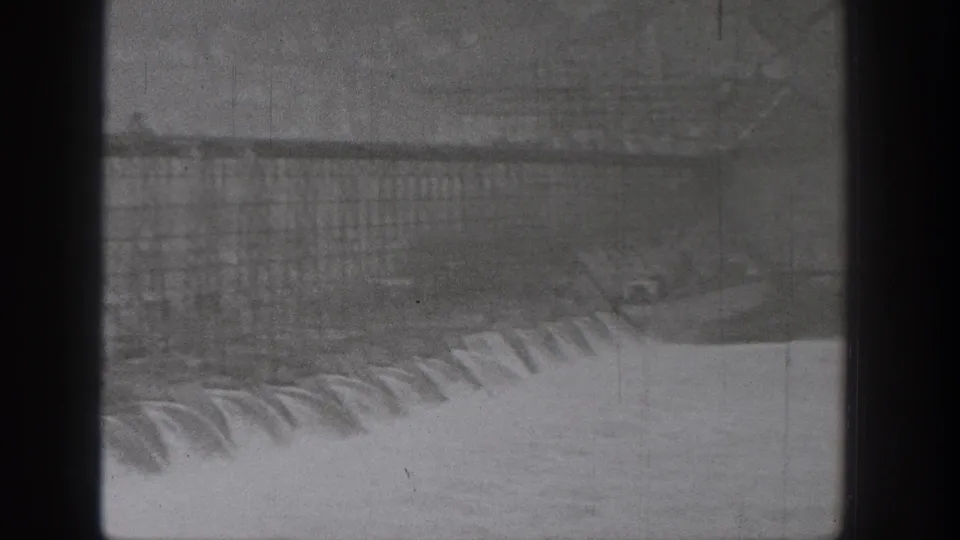1938 Washington dam footage showing spillway water over concrete, with distant hillside and dark vertical obstruction in the foreground.