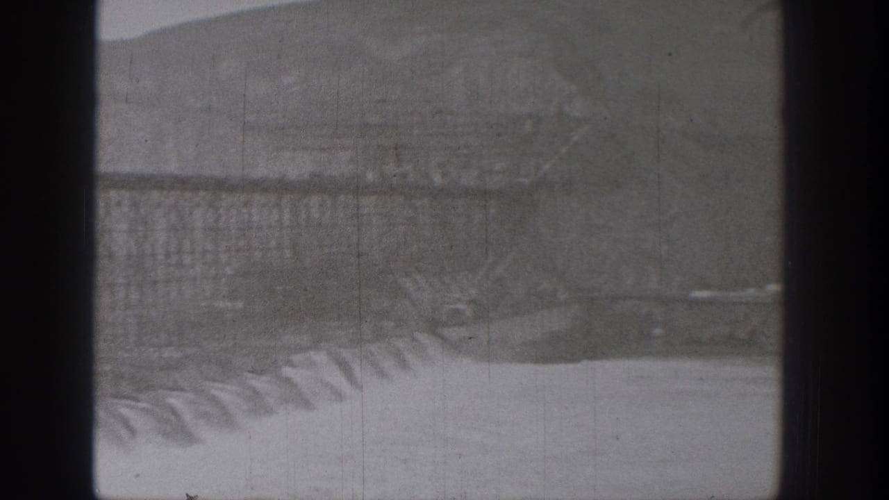 Black-and-white 8mm view of a Washington dam spillway, with water cascading over concrete and hills in the background.