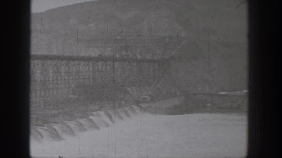 Black-and-white 8mm view of a Washington dam spillway, with water cascading over concrete and hills in the background.