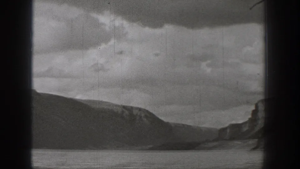 1938 Washington 8mm landscape frame with a broad lake, low mountain ridge, distant cliff, clouded sky, and visible film wear.