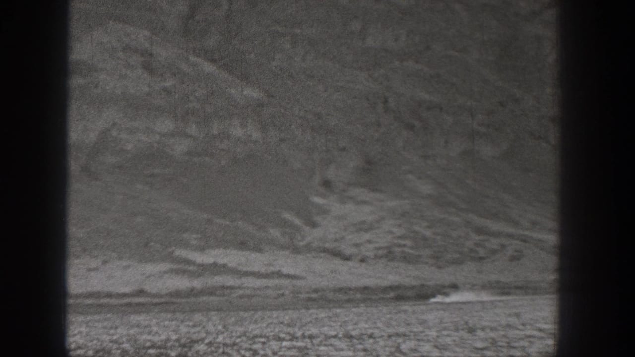 A distant vehicle appears at the base of rugged Washington desert hills, trailed by dust in grainy 8mm footage.