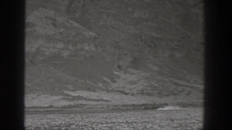 A distant vehicle appears at the base of rugged Washington desert hills, trailed by dust in grainy 8mm footage.