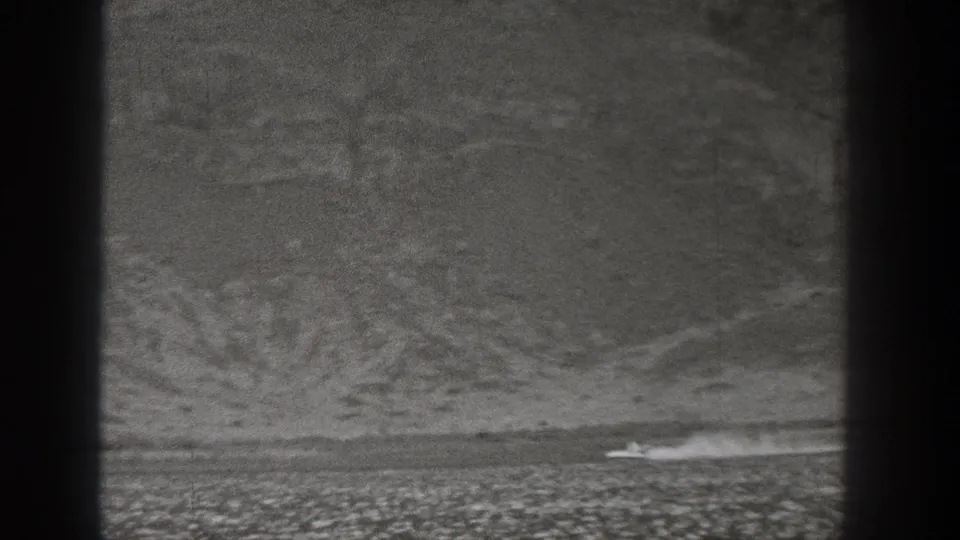 Grainy 1938 Washington frame with a small moving vehicle and dust trail against flat arid land and low hills.