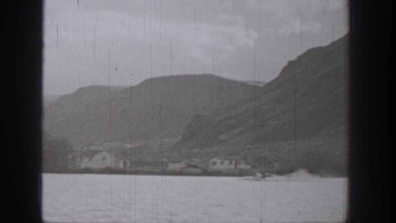1938 Washington 8mm frame showing a lake, hillside shoreline, small distant buildings, cloudy sky, and visible film gate edges.