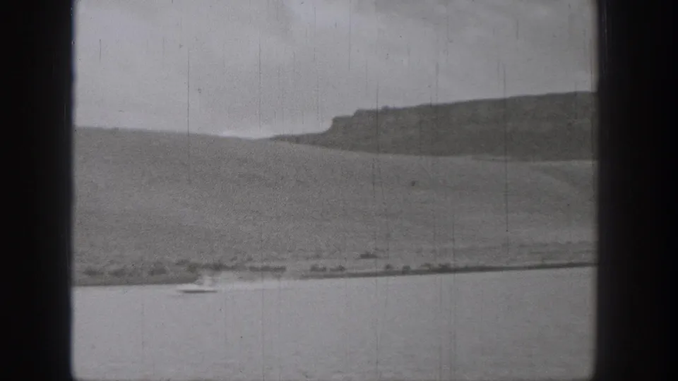 1938 Washington lakeside 8mm frame with open water, distant sloping hills, a low shoreline, and a small boat near the foreground edge.