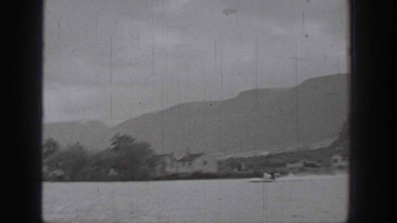 Wide black-and-white 8mm view of a Washington lake, with a small skier or boat in the foreground and layered mountains beyond the shore.