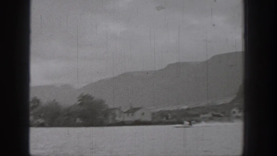 Wide black-and-white 8mm view of a Washington lake, with a small skier or boat in the foreground and layered mountains beyond the shore.