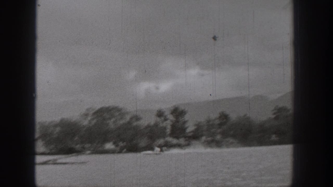 Soft 1938 Washington lake frame showing trees along the shore, mountains in the distance, and a small boat or skier near the waterline.