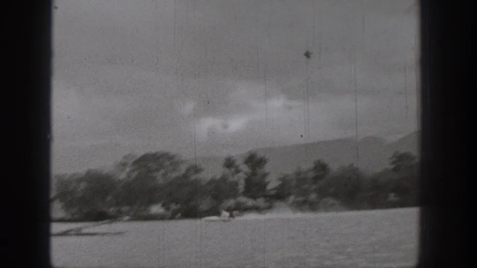 Soft 1938 Washington lake frame showing trees along the shore, mountains in the distance, and a small boat or skier near the waterline.