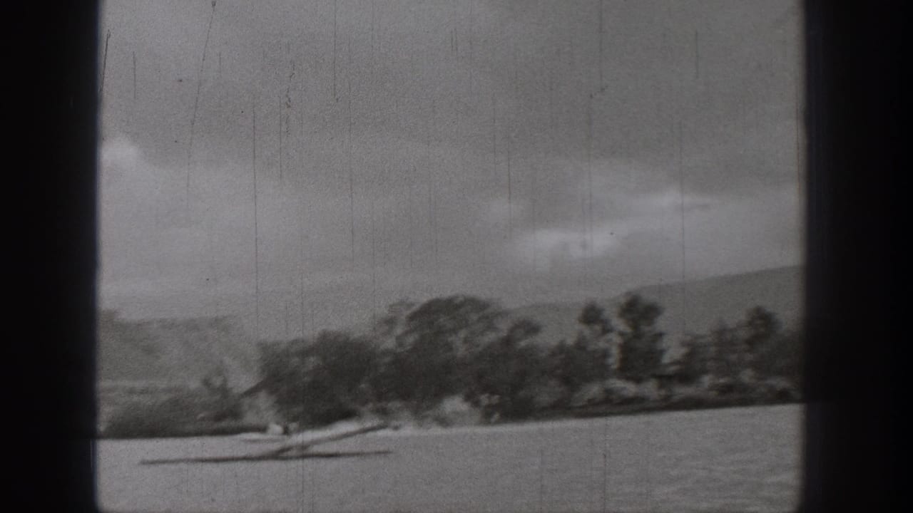 Black-and-white 1938 Washington 8mm frame showing a small boat on a lake with trees, shoreline, low hills, and overcast sky.