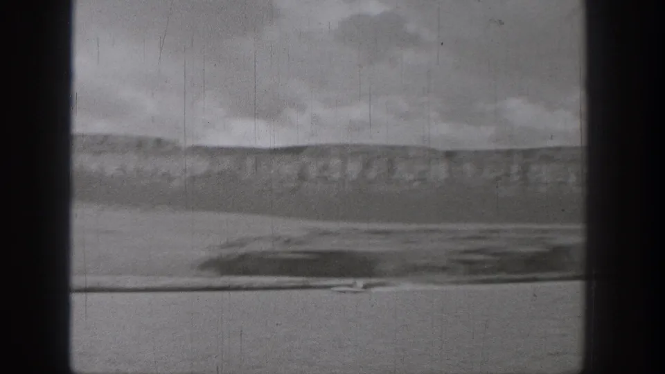 Black-and-white Washington lake view with a distant boat, low hills, and cloudy sky, framed by dark 8mm scan borders.