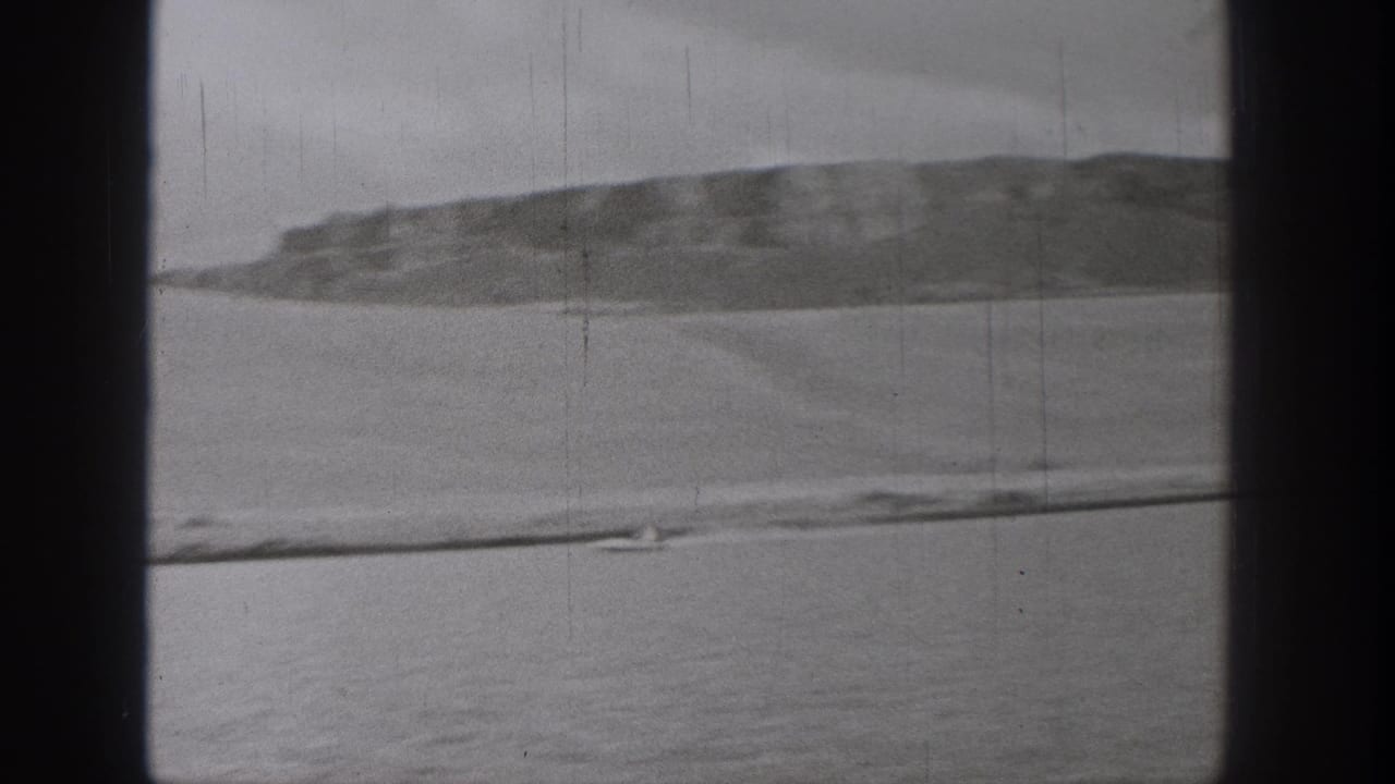 Black-and-white 8mm Washington lake frame showing a small boat low in frame, broad water, distant hills, and an overcast sky.