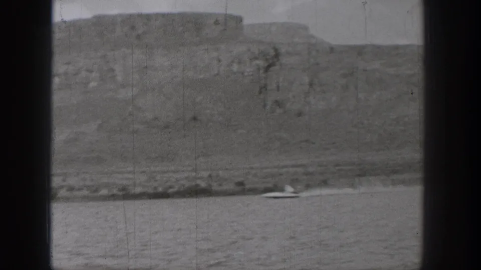 A small speedboat crosses the water near the shoreline below rugged Washington bluffs in grainy 1938 home-movie footage.
