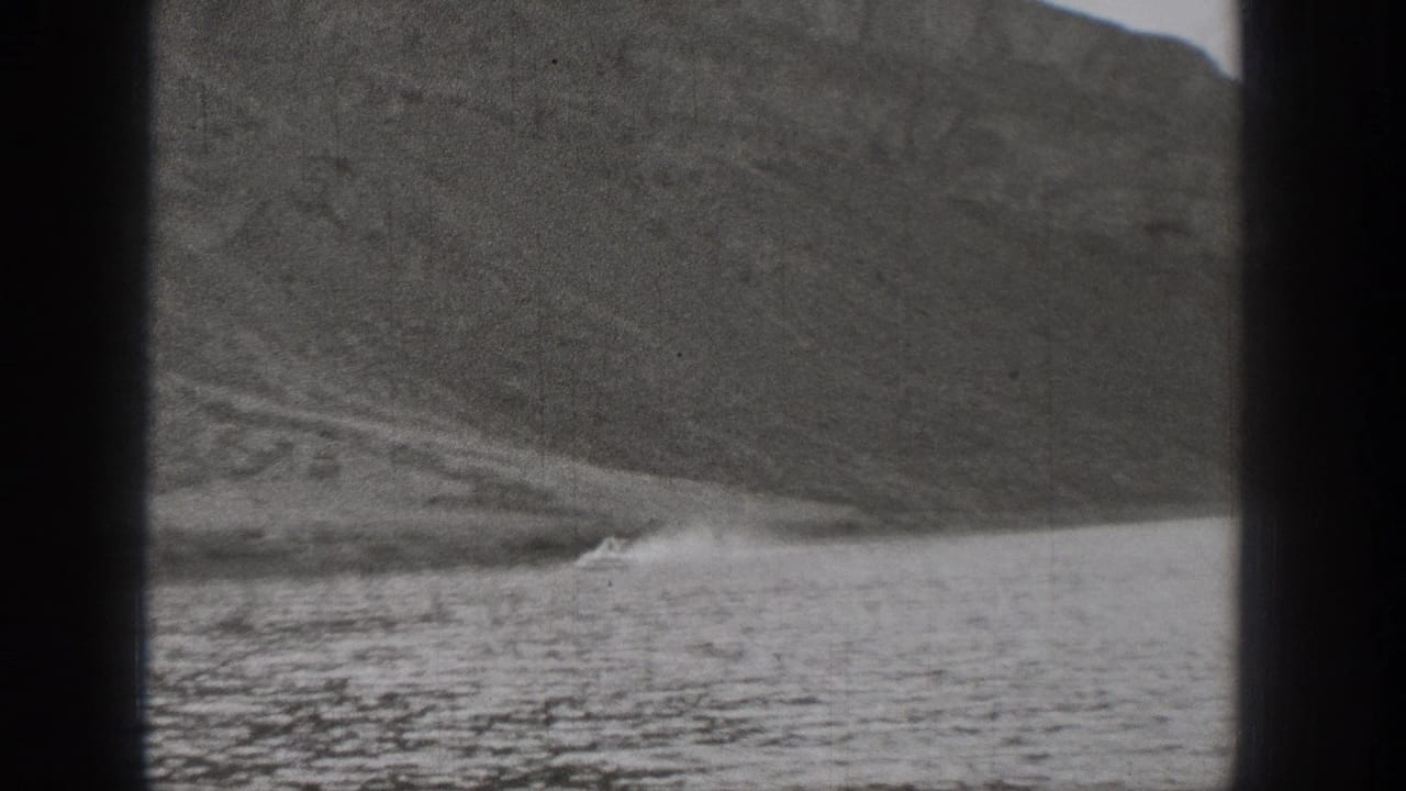 Grainy 1938 Washington 8mm frame showing water, shoreline, and a patch of white spray against a rocky mountain slope.
