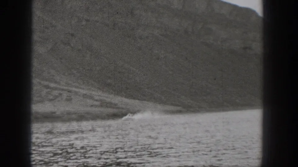 Grainy 1938 Washington 8mm frame showing water, shoreline, and a patch of white spray against a rocky mountain slope.