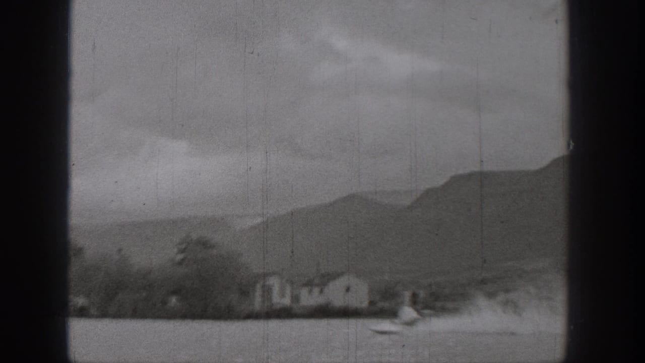 1938 Washington lake view with a small boat and wake near shoreline houses, trees, and low mountains.
