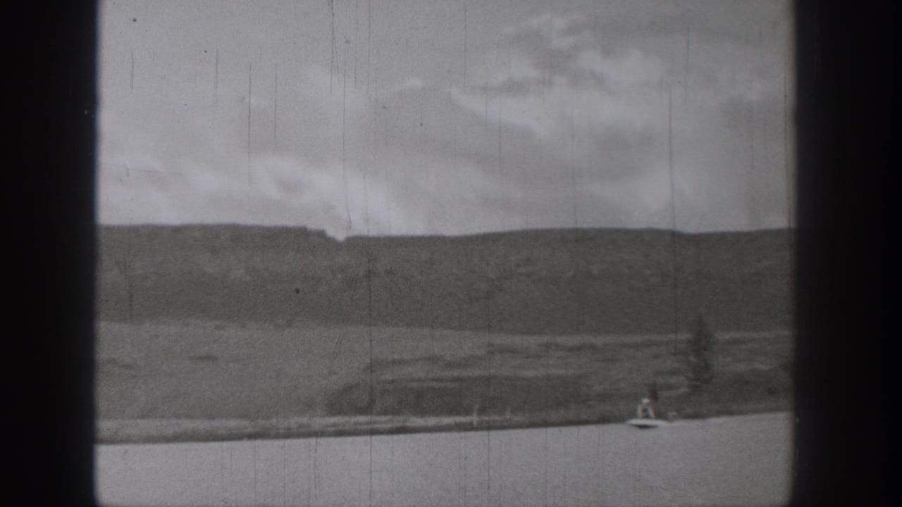 1938 Washington home-movie frame showing a distant rider on a motorized surf-like device on a calm lake with hills and cloudy sky.
