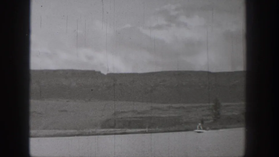 1938 Washington home-movie frame showing a distant rider on a motorized surf-like device on a calm lake with hills and cloudy sky.