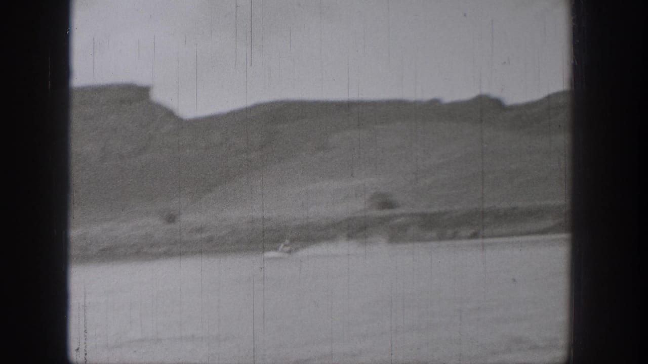 1938 Washington 8mm frame showing calm lake water, low shoreline, and distant rugged cliffs under a pale sky.