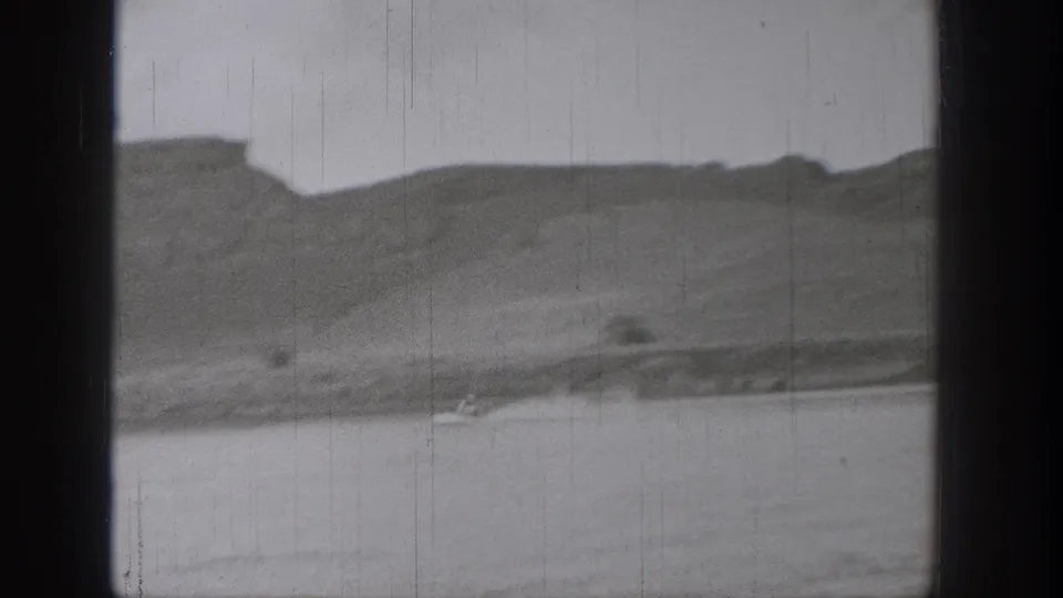 1938 Washington 8mm frame showing calm lake water, low shoreline, and distant rugged cliffs under a pale sky.