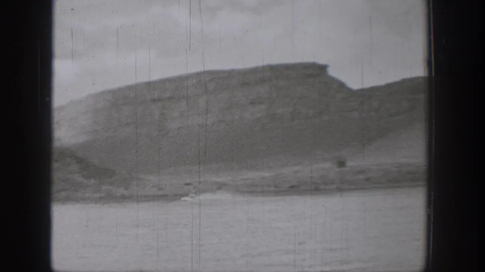 1938 Washington 8mm frame of a broad cliff ridge beyond calm lake water, with a small dark speck near the distant shore.