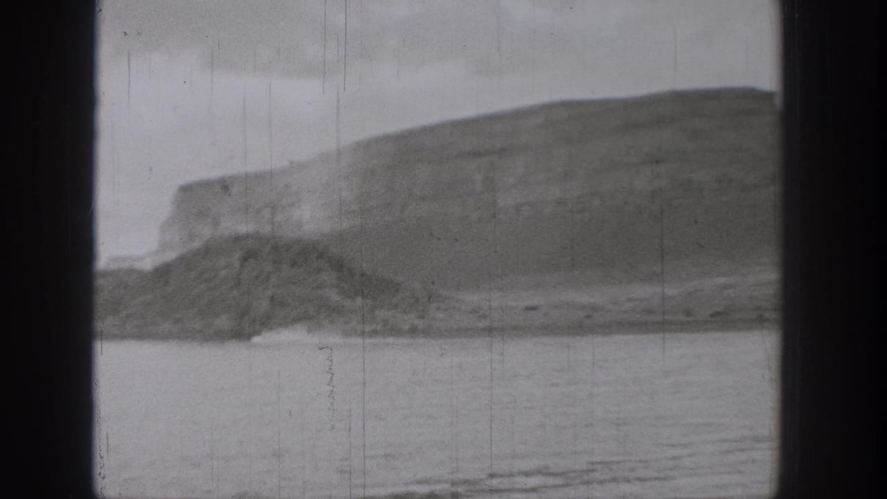1938 Washington 8mm frame showing a calm lake beneath a large layered cliff face and rounded hillside.
