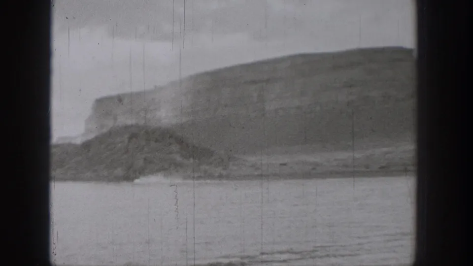 1938 Washington 8mm frame showing a calm lake beneath a large layered cliff face and rounded hillside.