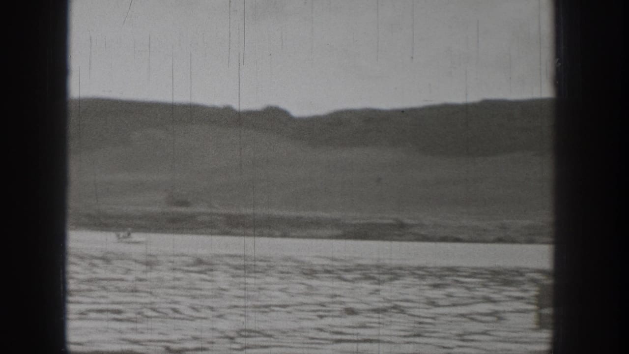 Monochrome home-movie frame showing Washington lakeside hills and textured water with visible archival film marks.