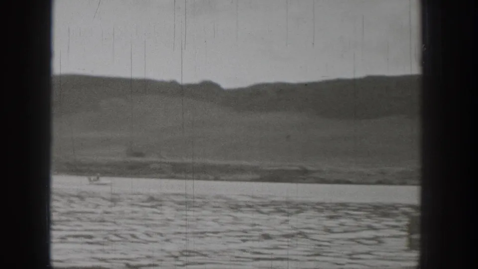 Monochrome home-movie frame showing Washington lakeside hills and textured water with visible archival film marks.