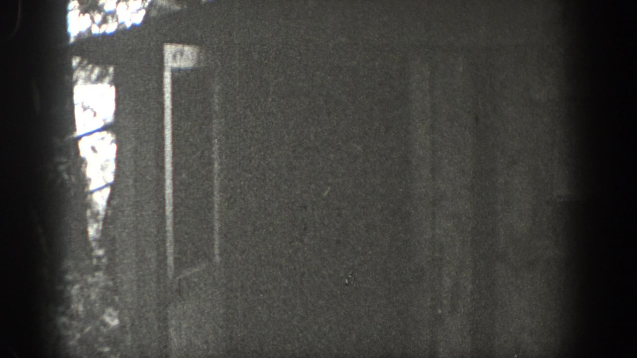 Washington 1938 home-movie frame showing a rustic cottage doorway in heavy grain, partly blocked by trees and dark film-edge exposure.