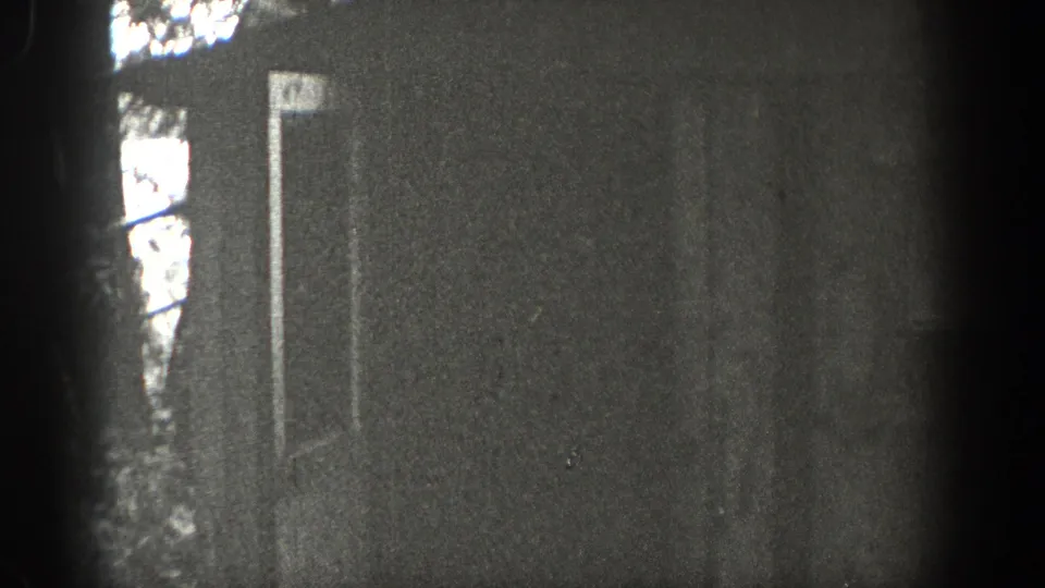 Washington 1938 home-movie frame showing a rustic cottage doorway in heavy grain, partly blocked by trees and dark film-edge exposure.