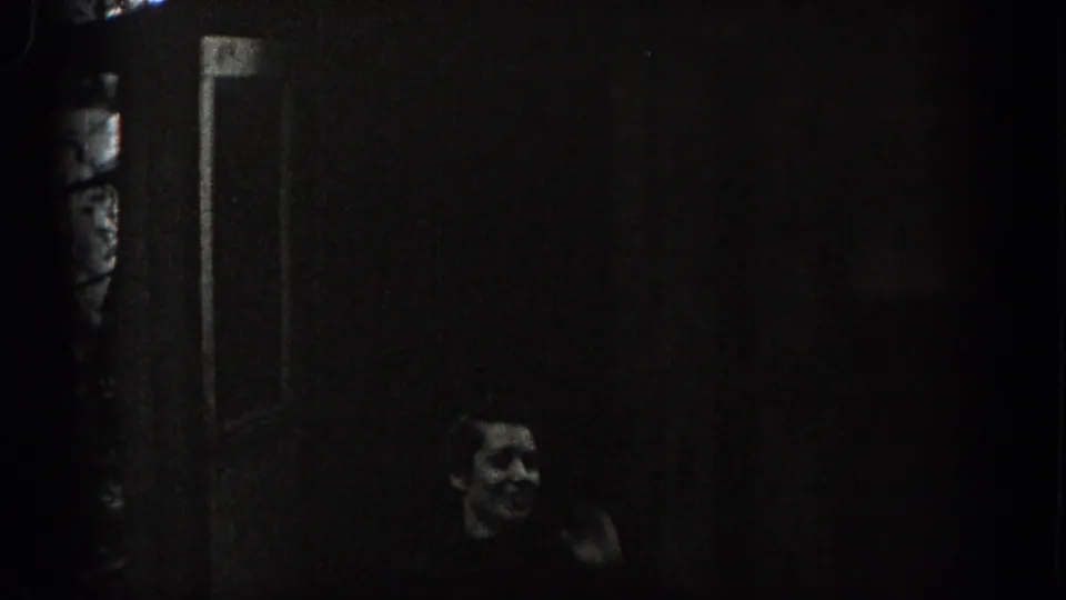 Washington 1938 home-movie frame showing a woman near a cottage doorway, her face faintly visible in low exposure.