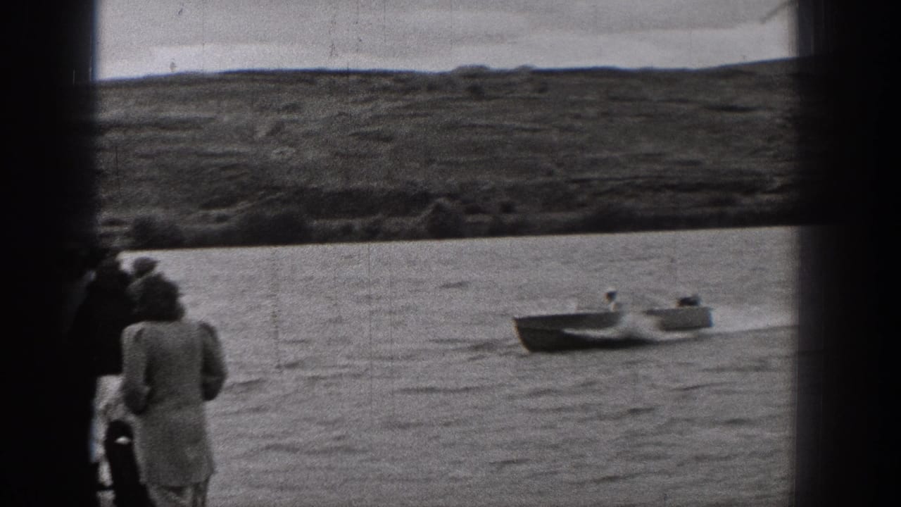 Home-movie frame from 1938 Washington showing distant shore viewers beside the water while a small open boat passes in the background.